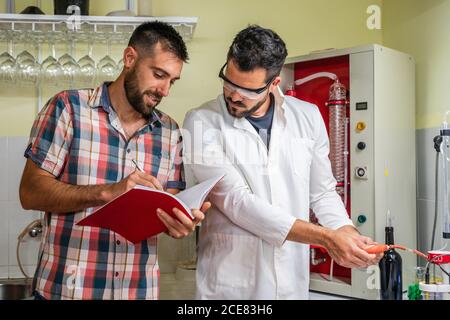 Adult man writing down information in copybook while male partner in uniform using wine measuring tool in laboratory near glassware Stock Photo