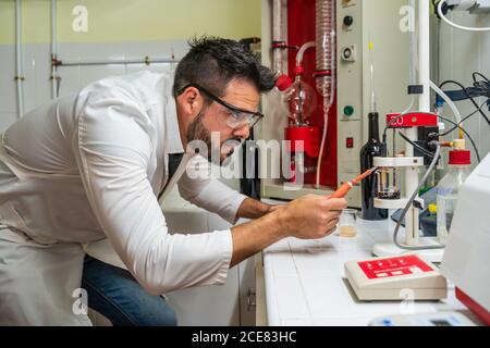 Side view of serious chemist in uniform and protective eyeglasses using transparent flask with red wine and measuring tools at work in lab Stock Photo