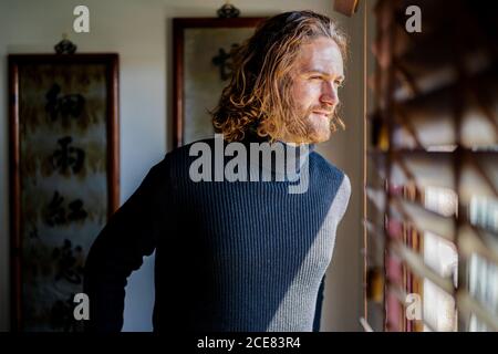 Middle aged male in casual wear and with long hair standing near window with shutters and observing street while chilling at home during weekend Stock Photo