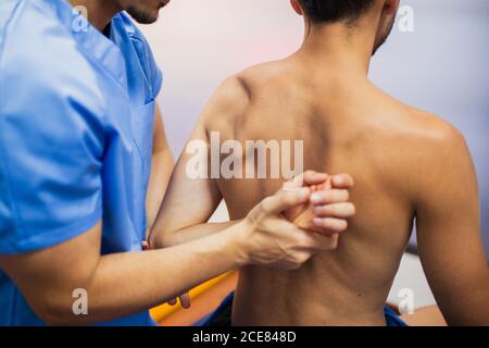 Crop of back view of orthopedist in uniform checking up shoulder joint of fit man sitting on examination couch with flexed arm in hospital and looking away Stock Photo