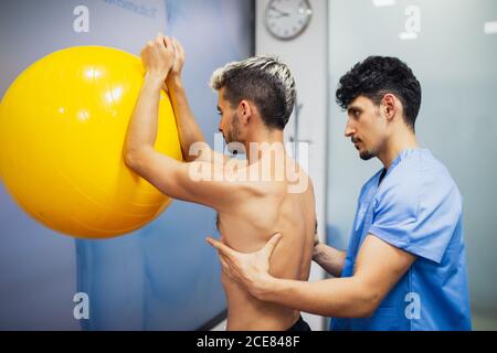 Side view of serious young osteopath in uniform supporting back of fit anonymous man putting elbows on colorful fitness ball during rehabilitation process in hospital Stock Photo