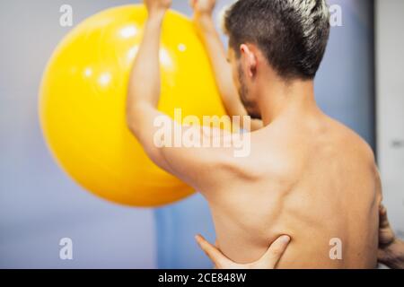 Crop of side view of young osteopath supporting back of fit anonymous man putting elbows on colorful fitness ball during rehabilitation process in hospital Stock Photo