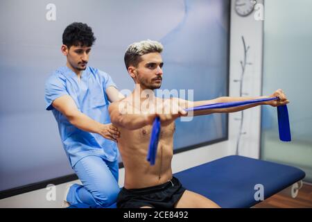 Sports medicine doctor checking up back of sportive man with dyed hair stretching elastic tape during rehabilitation period and looking away Stock Photo