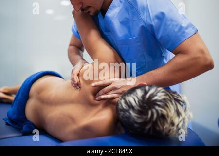 Osteopath in blue uniform examining back of unrecognizable slim male patient with dyed hair lying on examination table in clinic Stock Photo