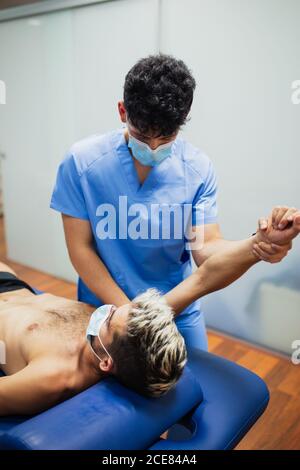 Osteopath with mask in blue uniform examining arm of slim male patient with mask and dyed hair lying on examination table in clinic Stock Photo