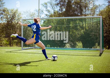 Side view of unrecognizable female athlete shooting ball into goal while playing football on field Stock Photo