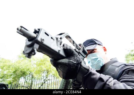 From below of serious spec ops police officer in  protective uniform and medical mask aiming submachine gun during police operation Stock Photo