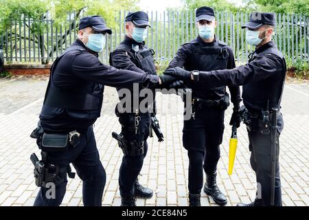 Serious anonymous police officers in protective gears and medical masks armed with guns and batons putting hands together while preparing for operation