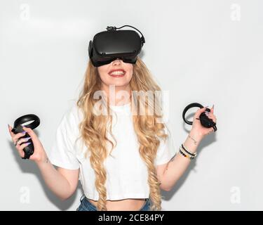 Delighted female wearing goggles of virtual reality playing video game with controllers while standing on white background in studio Stock Photo