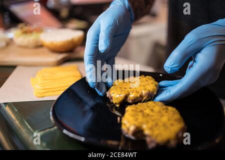 High angle of anonymous chef in medical gloves adding cheese on burger bun Stock Photo
