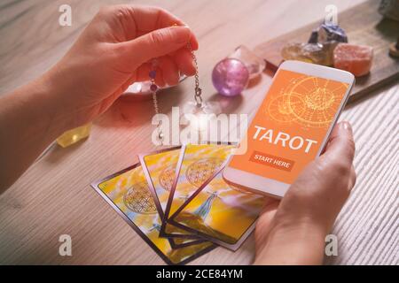 Tarot cards dowsing tool in hand and smartphone with modern fortunetelling application on screen as a concept of psychic advisor or newest ways of div Stock Photo