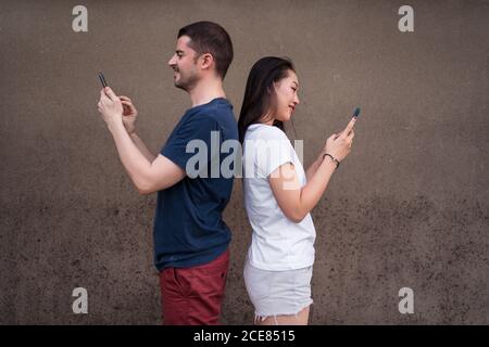 Side view of multiethnic man and Woman standing back to back and using smartphones to text messages in social media against gray wall Stock Photo