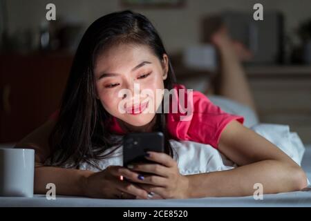 Positive ethnic Woman smiling and browsing dating app on smartphone while lying on bed in evening at home Stock Photo