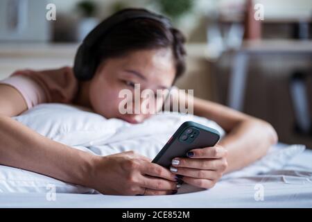 Unhappy ethnic Woman lying on bed and using smartphone in cozy room at home Stock Photo