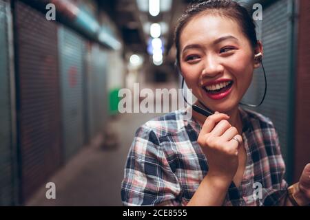 Joyful young Asian female student in casual clothes communicating with friends through headset while standing on blurred urban background Stock Photo