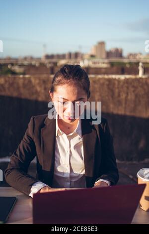 Front view of young Asian business lady in formal outfit using laptop while working remotely on rooftop terrace in city Stock Photo