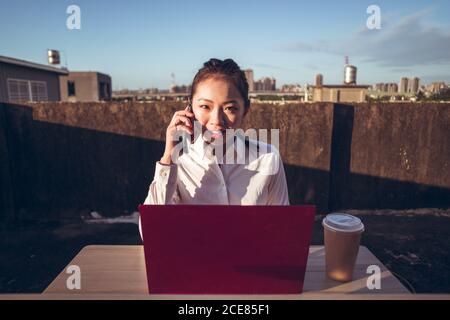 Front view of young Asian business lady in formal outfit talking on smartphone and using laptop while working remotely on rooftop terrace in city Stock Photo