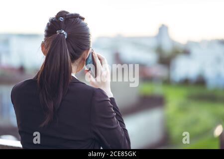 Back view of young Asian business lady in formal outfit talking on smartphone while working remotely on rooftop terrace in city Stock Photo