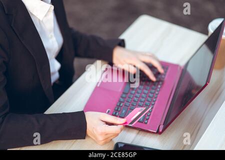 Crop of above view of young Asian business lady in formal suit with credit card using laptop during online payment while sitting at table at workplace on terrace Stock Photo