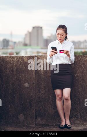 Serious young Asian businessWoman in formal wear holding credit card and smartphone with bank customer service while standing on rooftop of city building Stock Photo