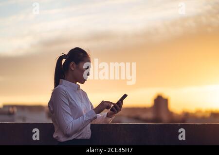 Side view of concentrated young Asian businessWoman in formal outfit using smartphone while standing on rooftop of city building at sunset Stock Photo