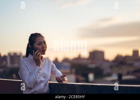 Side view of young Asian business lady in formal outfit talking on smartphone while working remotely on rooftop terrace in city Stock Photo