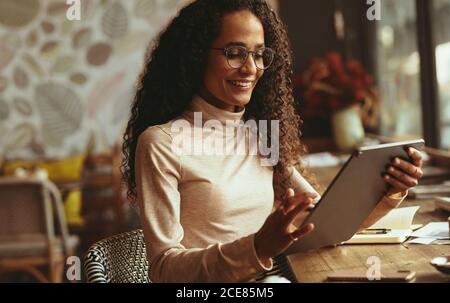 Woman sitting at cafe using her digital tablet. Smiling female in casuals working on her tablet pc from a coffee shop. Stock Photo