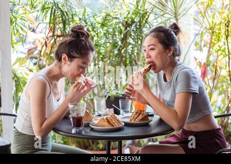 Young Asian female friends in summer wear relaxing at table in cafe and having conversation while eating delicious desserts Stock Photo