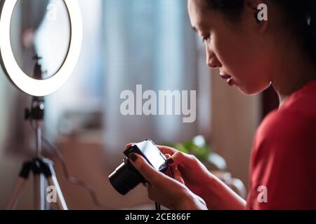 Side view of focused young Asian Woman sitting at table with cosmetics and tools for video shooting and checking video on camera while creating content for beauty blog at home Stock Photo