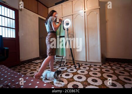 Low angle side view of young Asian female in stylish outfit standing in front of tripod with camera and ring light and demonstrating elegant dress while shooting video for fashion vlog at home with cute dog lying nearby Stock Photo