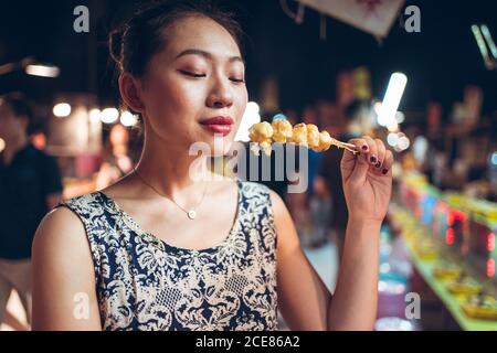 Content Asian female eating delicious fried quail eggs on stick while standing in Zhubei Night Market Stock Photo