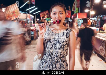 Content ethnic female eating sugar coated cherry tomato from wooden stick while standing in Zhubei Night Market looking at camera Stock Photo