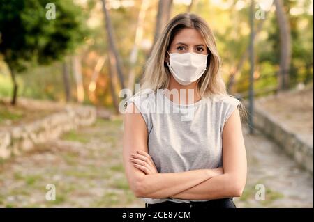 Young female in casual clothes and protective mask for coronavirus prevention looking at camera while standing on alley in green park Stock Photo