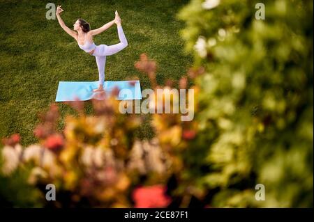 From above side view of slim female in sportswear balancing on leg in Natarajasana while practicing yoga on green lawn in park Stock Photo