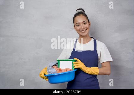 Housekeeping service. Happy maid woman in uniform holding basket with laundry and green plastic box with washing capsules, smiling at camera while standing against grey wall. Cleaning, housework Stock Photo