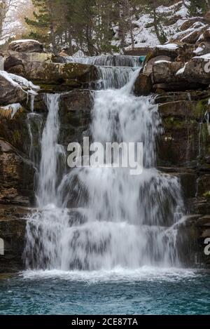 Picturesque landscape of small waterfall and pool with turquoise water located among boulders covered with snow against green coniferous forest Stock Photo