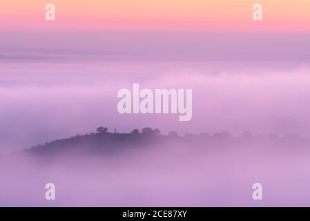 Drone view of hilly terrain covered with green trees under colorful pink clouds and fog with distant anonymous person silhouette in Toledo Stock Photo