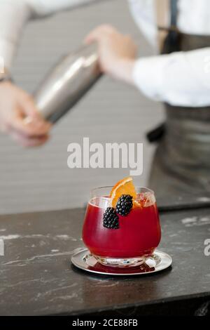 Glass of red refreshing cocktail with orange slice and blackberries placed on coaster on bar with blurred bartender on background Stock Photo