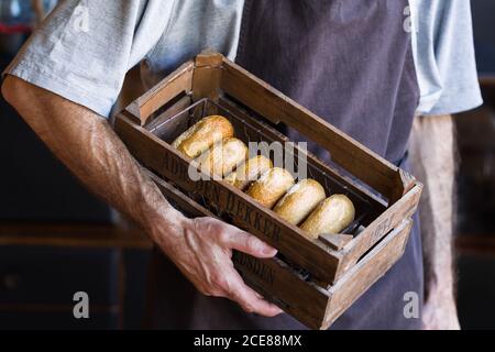 From above of crop unrecognizable male baker in apron standing with wooden box full of tasty freshly baked doughnuts with golden surface and tender texture Stock Photo