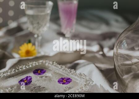 Above view of cocktail decorated with purple flower petals served on white tablecloth with vintage styled glasses and bottle on sunny day Stock Photo