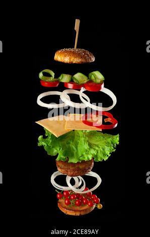 levitating cheeseburger ingredients: juicy meat cutlet, cheese, sesame bun, lettuce, white onion rings, slice of tomato, cucumber on black background. Stock Photo