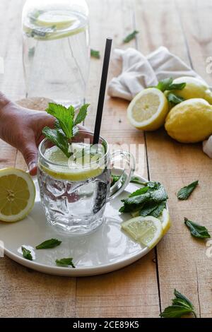 Crop unrecognizable person taking glass cup of tasty cold lemonade with soda water and mint leaves placed on wooden table Stock Photo