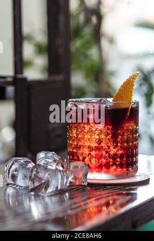 Melting cubes of ice placed on table near glass cup of alcohol beverage with orange peel in bar Stock Photo