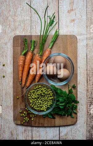 Top view of ripe carrots and basil arranged with canned peas and boiled eggs on wooden chopping board in kitchen Stock Photo