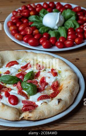 Round dish with yummy Pizza Margherita decorated with basil and dish with small oval tomatoes and basil placed on wooden table in Italian pizzeria Stock Photo