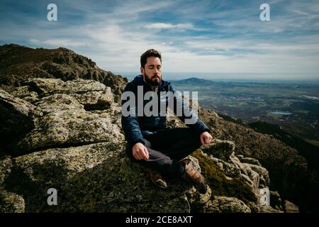 Bearded male traveler sitting cross legged on rock and meditating with closed eyes against cloudy sky in Puerto de la Morcuera mountains in Spain Stock Photo