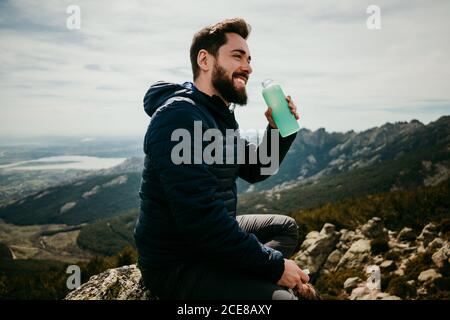 Side view of cheerful bearded man smiling and drinking fresh water while sitting on boulder against overcast sky in Puerto de la Morcuera mountains in Spain Stock Photo