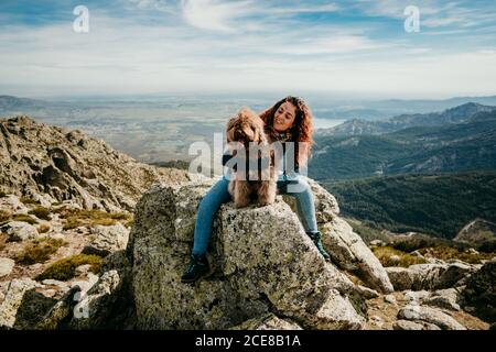 Full length female owner embracing fluffy Labradoodle while sitting on stone against cloudy sky in Puerto de la Morcuera mountains in Spain