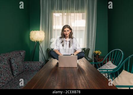 Full length of positive young female in casual outfit using laptop while sitting on top of wooden table in cozy creative apartment Stock Photo