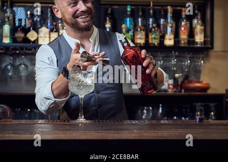 Professional happy young barman pouring alcohol cocktail from jigger into a glass on counter making beverage working in bar looking away Stock Photo
